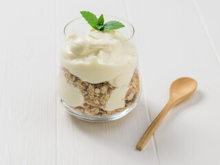 A large glass glass with granola and yogurt on a white table with a spoon.