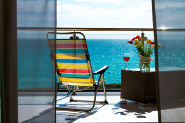 Multi colored striped beach chair, chaise lounge, a table with a glass of wine and flowers standing on the balcony on background of the seascape of blue calm sea and sunny clear sky.