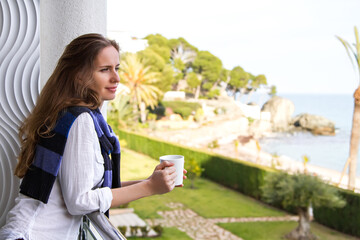 Beautiful young romantic long-haired woman with a mug in her hands, standing by the panoramic window on the balcony and thinking looking at the seascape.
