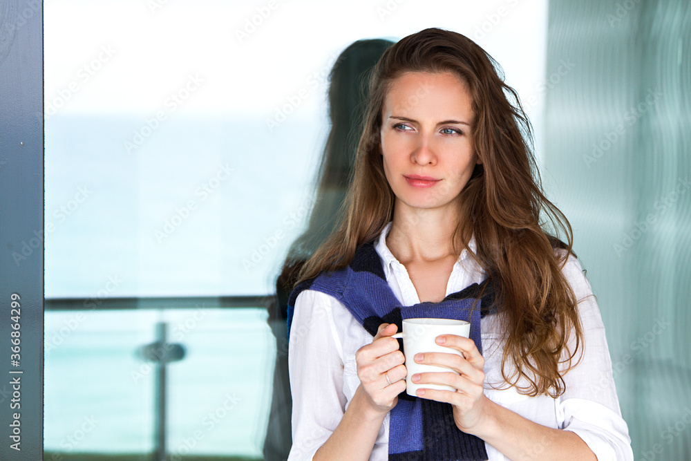 Wall mural beautiful young romantic long-haired woman with a mug in her hands, standing by the panoramic window