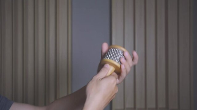 Caucasian adult male hands playing cabasa percussion, steel ball chain wrapped around wide cylinder, in recording studio with wall in background, close up static