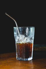 Half full glass with cola and a metal straw served on wood and black background 