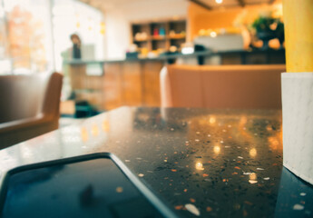Modern blurred coffee shop Scene. Phone and coffee cup on table as foreground. Soft blurred sofa chair, bar / counter shelves and window front in the background. Warm yellow red color theme.