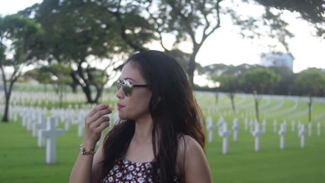 Asian woman with sunglasses walking through cemetery eating potato chips. Follow shot