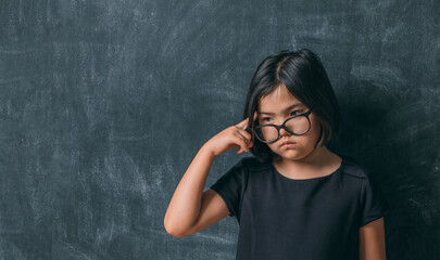 Back to school. Serious little girl in glasses thinking touching her head with a finger near blackboard. Child from elementary school. Education.