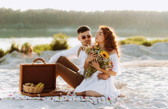 Handsome guy with glasses sitting near to a cute girl that holding a bouquet of wildflowers in her hands. Picnic at sunset.