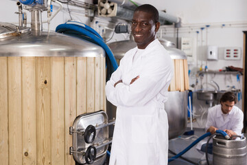 African american man brewer is standing near reservoir for beer in the factory during work