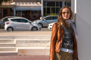 Obraz premium Portrait of a modern fashion beautiful woman with long hair and in sunglasses, brown jacket, smiling and looking at camera, on the background of a sunny cityscape view.