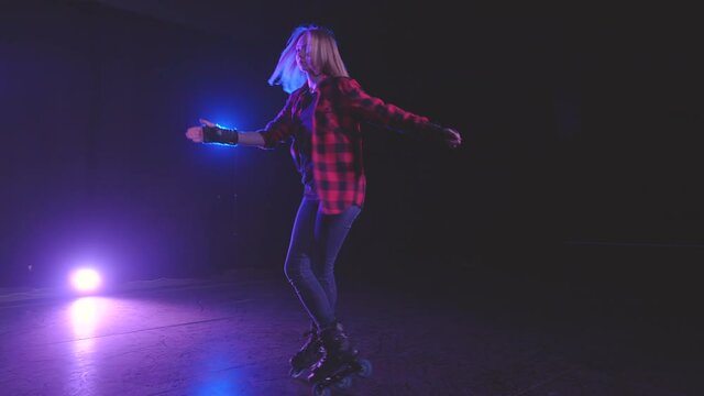 Close-up panoramic view of beautiful young roller skater woman, technically spinning on front wheels in one place under light of spotlights on dark stage in slow motion. Performance by active girl.