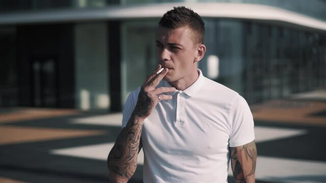 Young tattooed man in white polo shirt smokes cigarette in street, slow motion