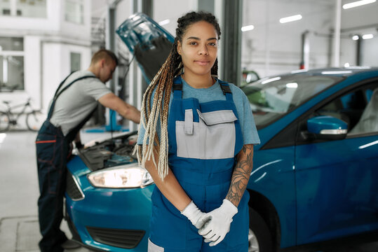 Smart service. Young african american woman, professional female mechanic looking at camera, while her male colleague repairing car in the background at auto repair shop