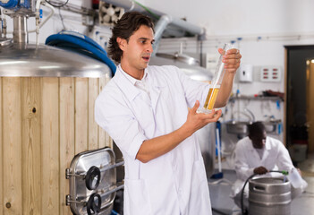 Young man expert is standing with flask for beer and analizying it in beer factory