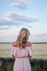 Attractive and beautiful girl in a pink dress in a wheat field. We are traveling together. Young lonely woman stands with her back in a field at sunset.