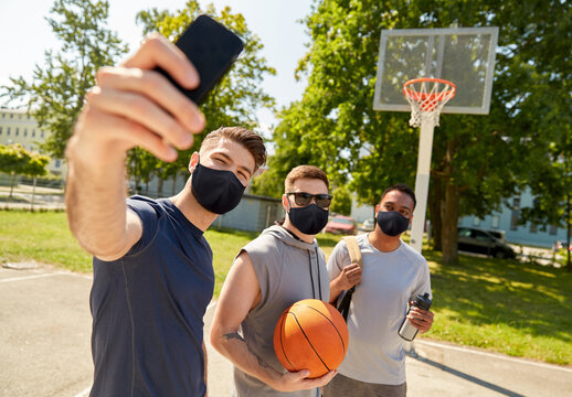 sport, leisure games and male friendship concept - group of happy men or friends wearing face protective masks for protection from virus disease taking selfie on outdoor basketball playground