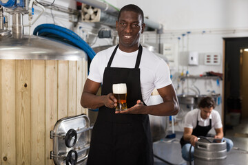 Fototapeta na wymiar African american man brewer in apron standing with glass of beer in brew-house, man on backround