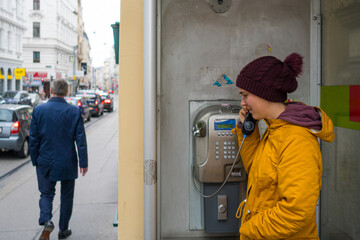 A young woman standing in front of an old payphone while calling someone. The street is right next to her with parked cars on the sidewalk.