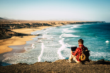 Travel and technology digital job nomad lifestyle people - man sitting on the coast work with laptop computer - wonderful beach in background - freedom from office life
