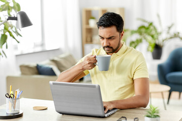 technology, remote job and lifestyle concept - indian man with laptop computer drinking coffee or tea at home office