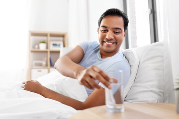 people, bedtime and rest concept - happy smiling indian man lying in bed at home and taking glass of water from table