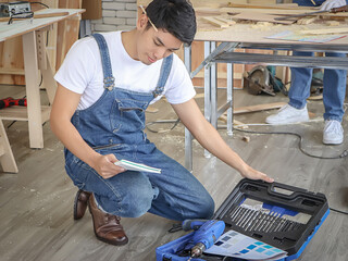 Young and handsome Asian repairman sitting  in indoor  workshop , reading instructiom with drill...