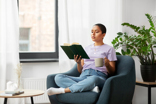 people and leisure concept - african american woman in glasses sitting on sofa and reading book at home