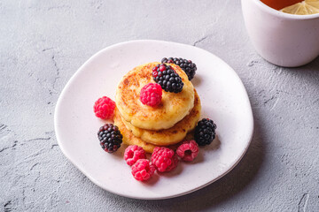Cottage cheese pancakes, curd fritters dessert with raspberry and blackberry berries in plate near to hot tea cup with lemon slice on stone concrete background, angle view