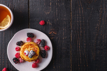 Cottage cheese pancakes, curd fritters dessert with raspberry and blackberry berries in plate near to hot tea cup with lemon slice on dark black wooden background, top view copy space