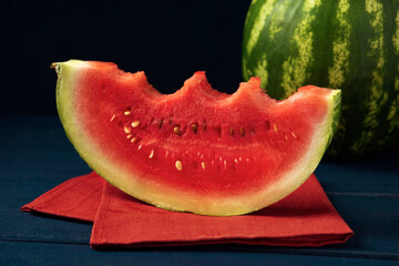 Bitten slice of watermelon with whole watermelon fruit on red napkin and dark blue wooden table.