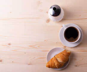 a white ceramic cup with black espresso, french croissant as breakfast or snack concept on rustic, natural texture wooden surface