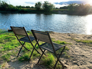 Beach chairs by the river. Evening sunset, sunshine. Natural landscape, river bank at sunset. Landscape, reflection in water, sun rays, grass on the shore.