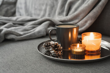 Aroma candles of orange color, coffee in a black mug and decorative pine cones served on a metal tray. Autumn or winter atmosphere
