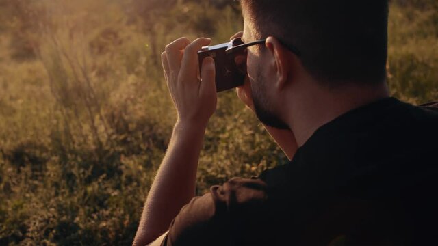 Close-up of a young man taking pictures in nature with a retro camera. Video from behind. Charismatic young man sets the camera