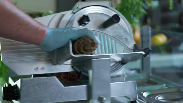 professional chef in gloves uses slicer and slices meat in kitchen of cafe or restaurant