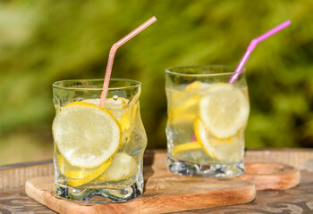 Lemonade in glass with slices of lemon and straws in the garden on sunlight, on a natural green background. Picnic with refreshing summer drinks.