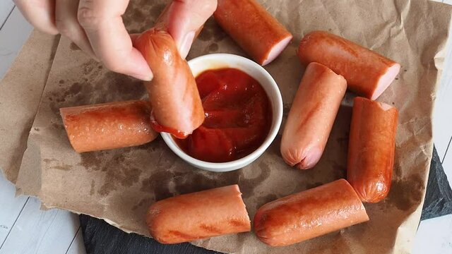 A girl dips a grilled sausage in tomato ketchup sauce.