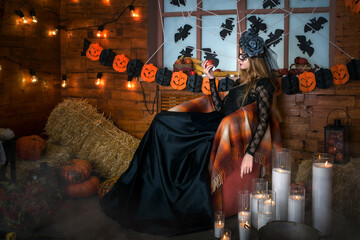 Halloween young beautiful witch is sitting in a chair and holding an apple in her hand. Candles are burning on the floor. Magic picture