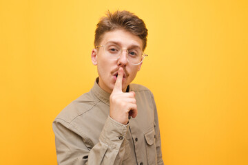 Close up portrait of a funny guy with mustache and glasses on a yellow background, shows a sign of silence and looks intently at the camera.Nerd put his finger to his lips and shows a gesture of thh