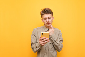 Thoughtful young man with mustache and glasses uses a smartphone against a yellow background,looks at the screen and thinks,wearing a shirt.Funny nerd with a serious face is using the phone. Isolated