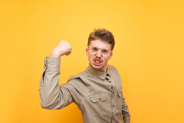 Portrait of emotional young man in glasses and shirt against yellow background, aggressively looks into camera and shows biceps. Expressive nerd looks into the camera with a crazy face and shouts.