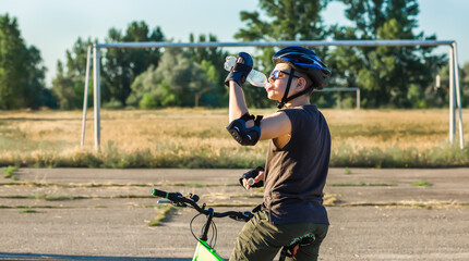A boy in a safety helmet on a bicycle drinks water from a plastic bottle. Thirst. Cycling. Sports stadium. Active lifestyle concept