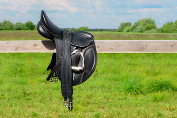 The black saddle and its girth is lying on the wooden hitching post in outdoors.