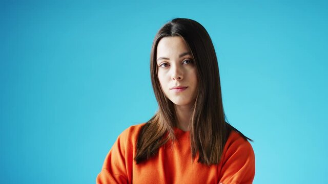 Studio portrait of confident young woman looking into camera with neutral expression and folded arms against blue background - shot in slow motion