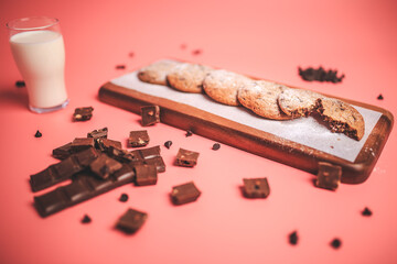 Freshly made Chocolate Chip Cookies on a wooden serving board. Cookies with a glass of fresh milk on the side along with chocolate chips and pieces of chocolate around on a pink background.Freshly mad