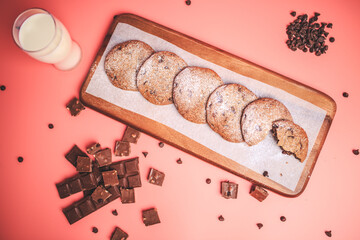 Freshly made Chocolate Chip Cookies on a wooden serving board. Cookies with a glass of fresh milk on the side along with chocolate chips and pieces of chocolate around on a pink background.Freshly mad