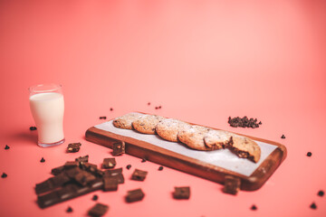 Freshly made Chocolate Chip Cookies on a wooden serving board. Cookies with a glass of fresh milk on the side along with chocolate chips and pieces of chocolate around on a pink background.Freshly mad