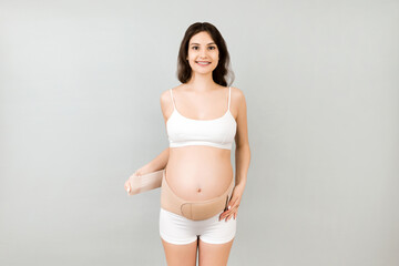 Fototapeta premium Portrait of pregnant woman in underwear dressing orthopedic bandage to reduce pain in the back at gray background with copy space. Orthopedic abdominal support belt concept