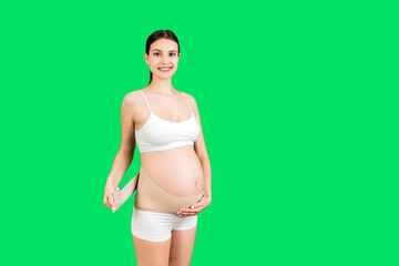 Portrait of pregnant woman in underwear dressing pregnancy corset on the third trimester at green background with copy space. Orthopedic abdominal support belt concept