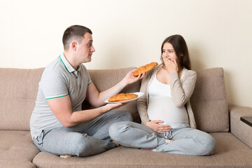 Fototapeta na wymiar Husband offers croissants to his pregnant wife but she refuses and makes stop gesture because she feels sick. Feeling bad during pregnancy concept