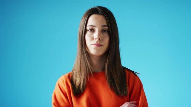 Studio portrait of confident young woman looking into camera with neutral expression and folded arms against blue background - shot in slow motion