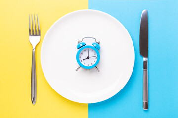 Blue alarm clock, fork, knife and empty plate on colored paper background. Intermittent fasting concept - 366919237